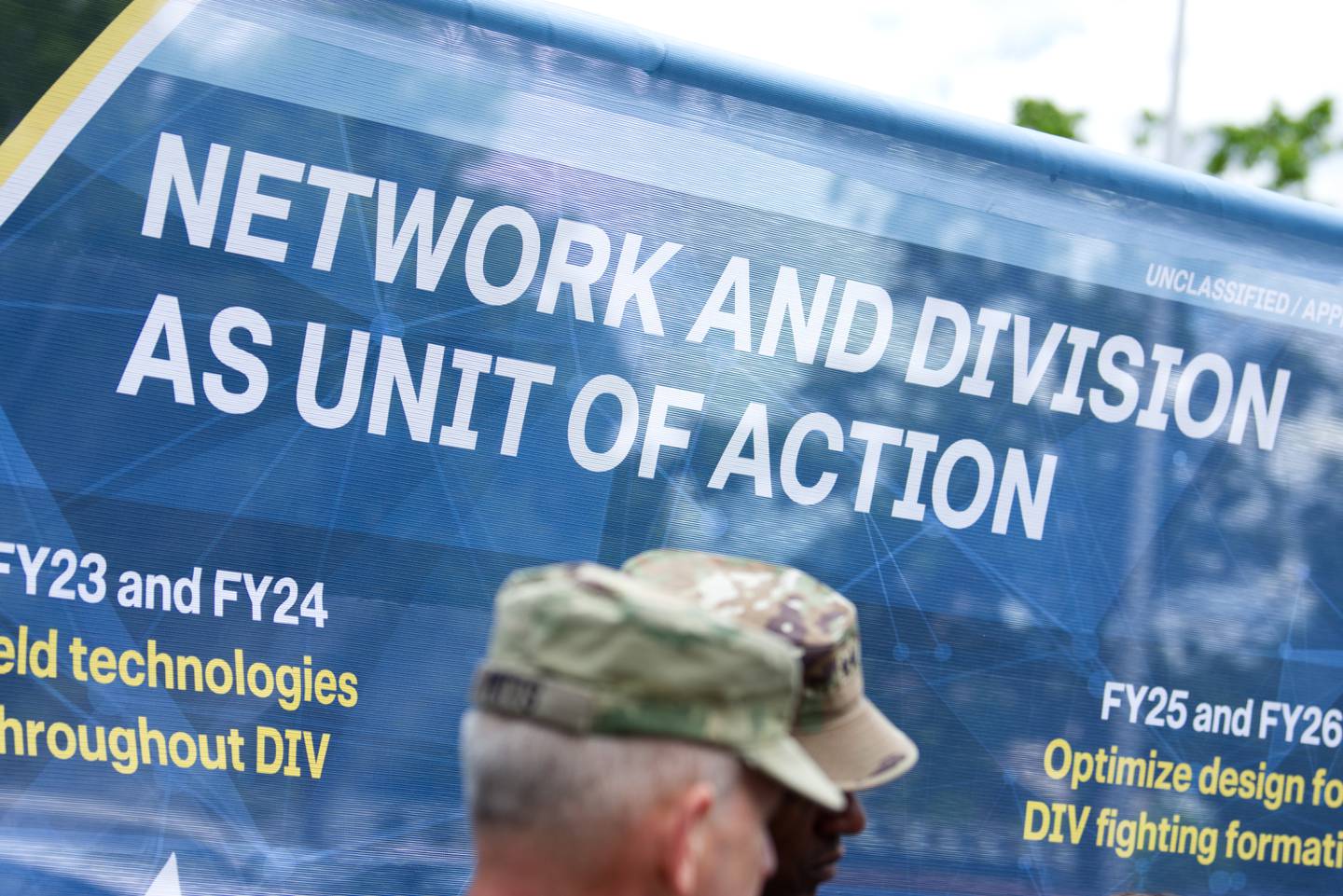 A banner set up for a tech demonstration at Fort Myer, Virginia, in May 2023 reads: "Network and division as unit of action." Two Army generals are seen in the foreground.